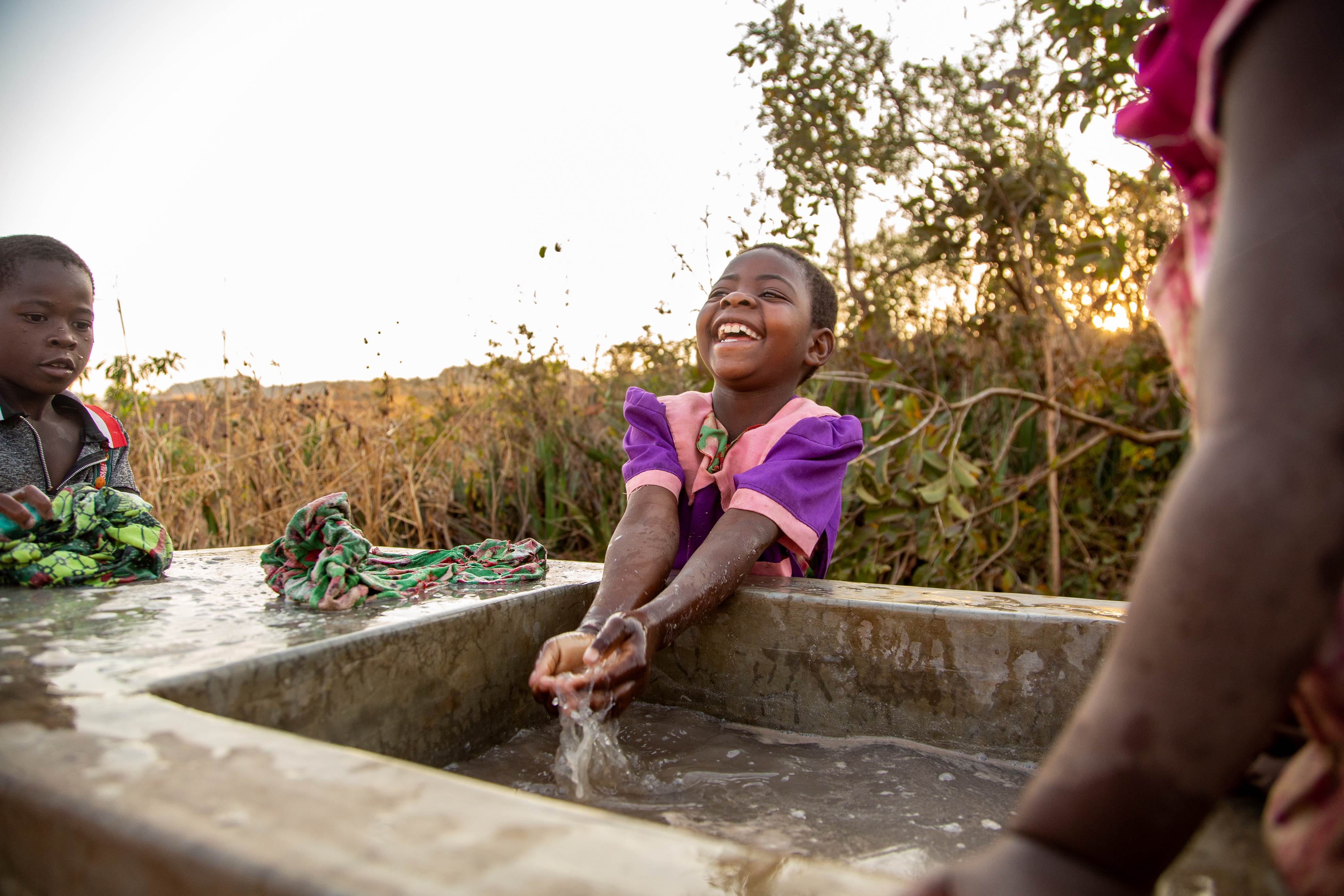 Girl from Malawi in bright purple dress laughing as she plays with the water from a local well