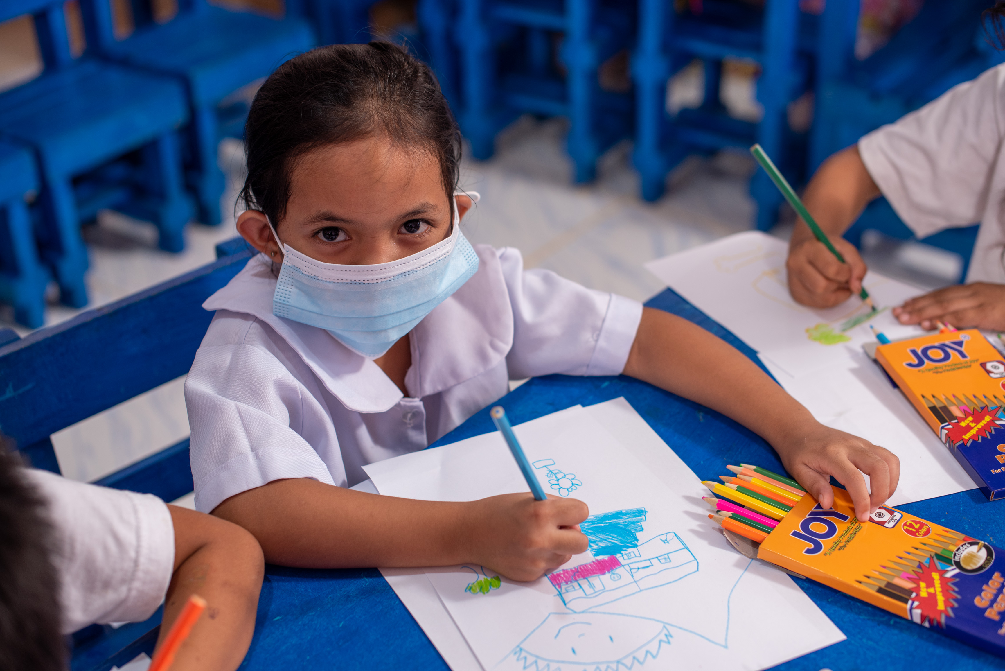 Girls from the Philippines wearing a face mask and school uniform looks up to the camera while drawing at school