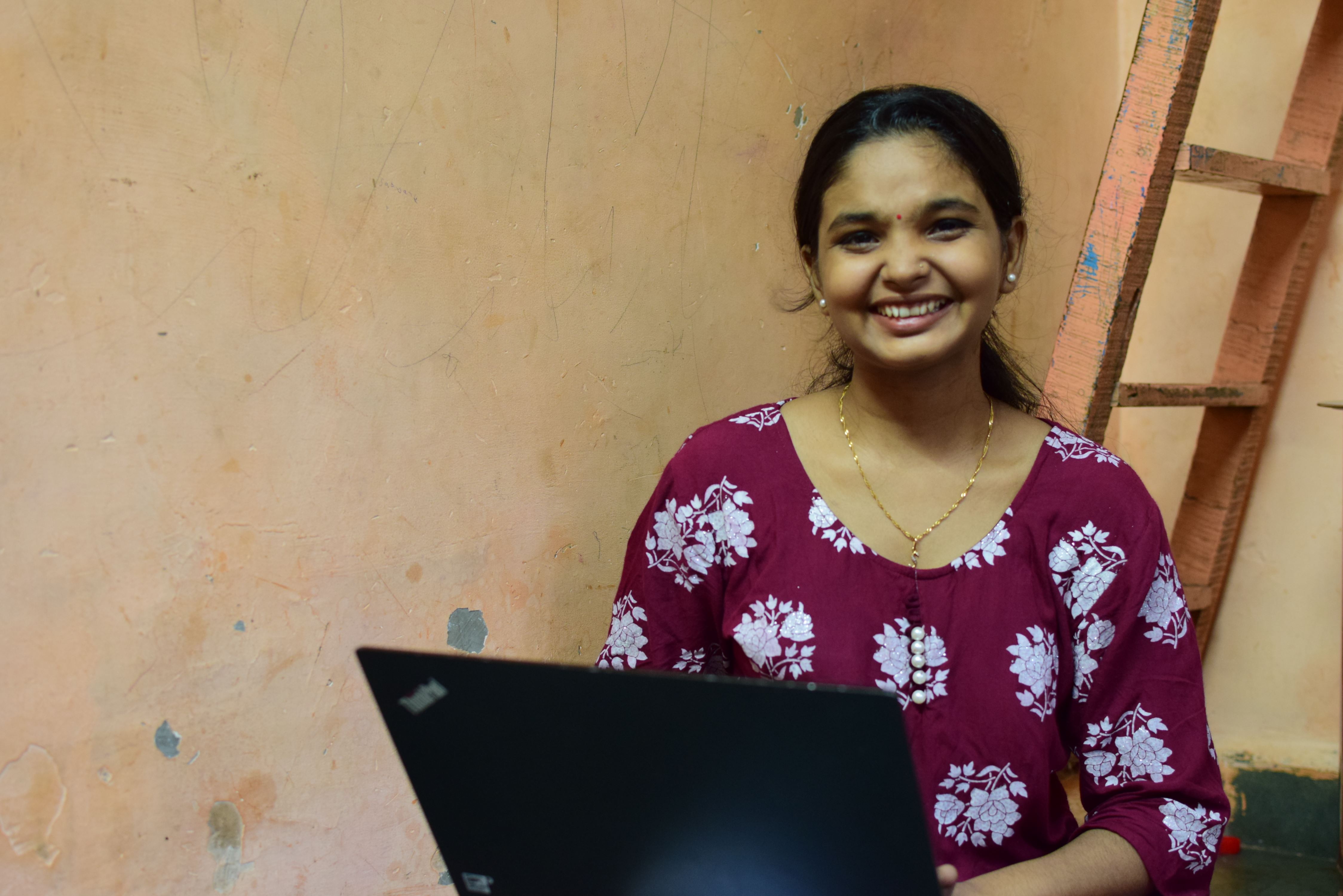Previously sponsored girl by World Vision smiling and looking at the camera in her home in India while working on her laptop