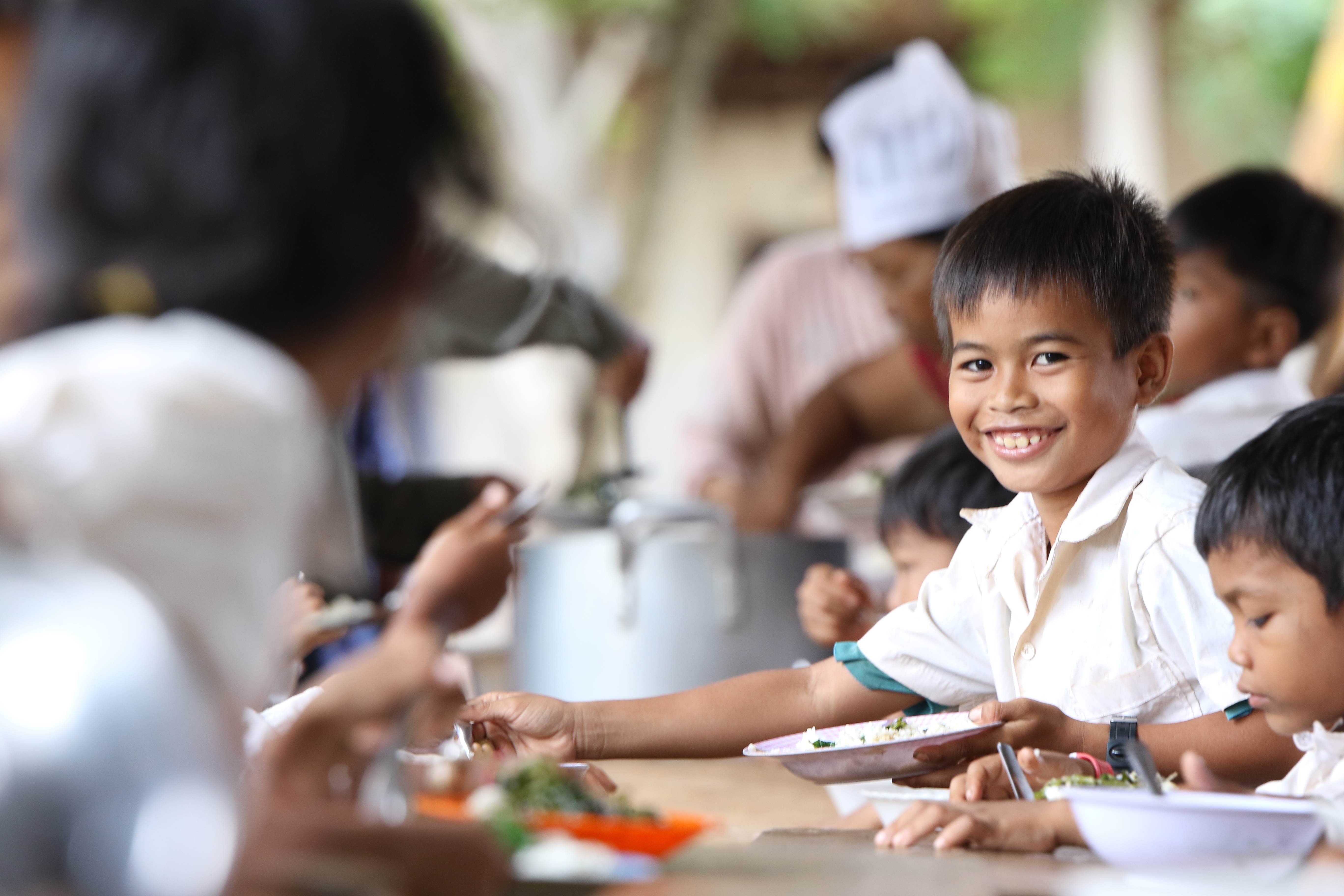 Boy smiles at the camera, surrounded by his schoolmates eating their free school meal in Cambodia