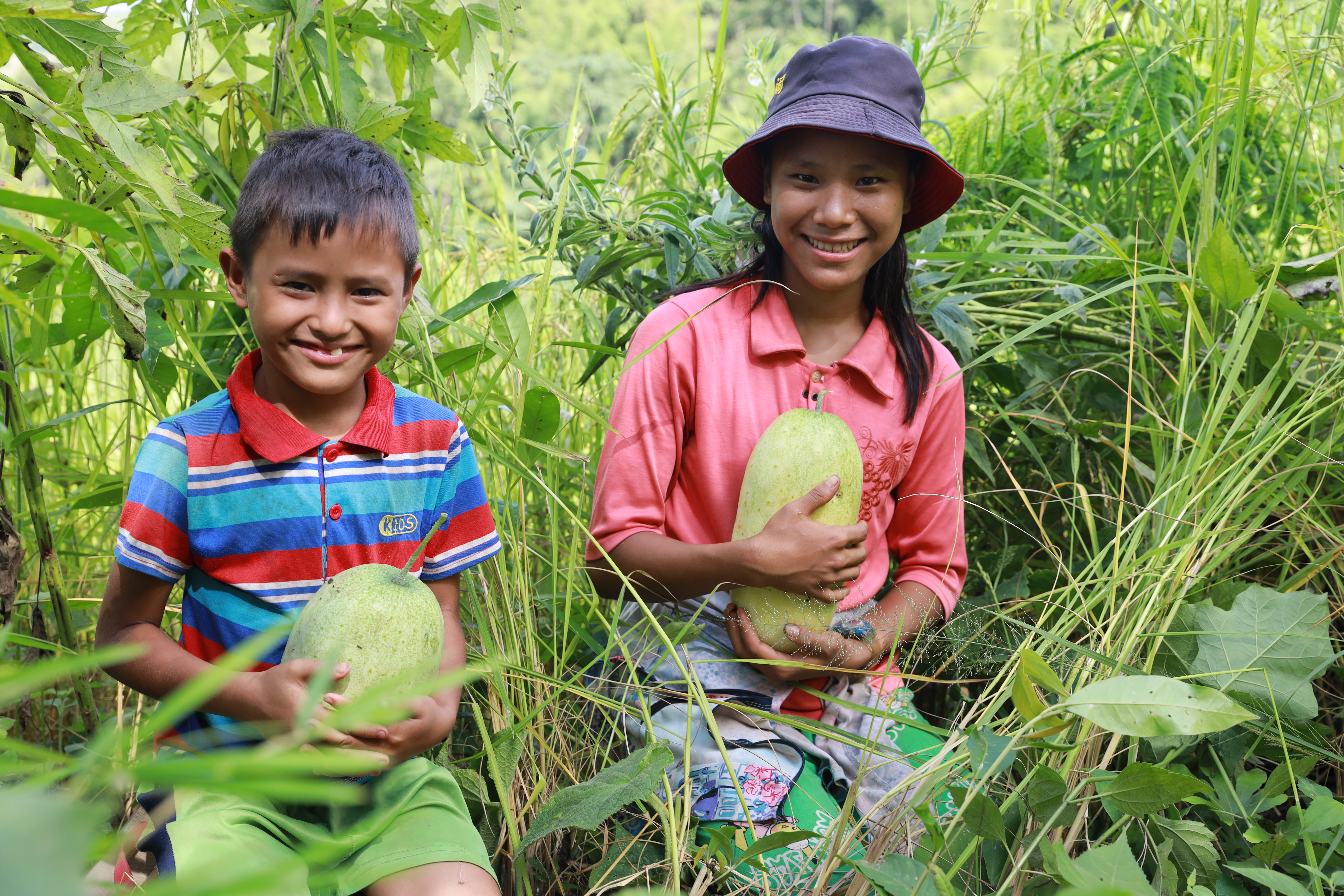 Brother and sister from Myanmar holding melons and smiling at the camera while standing in a green field of crops.