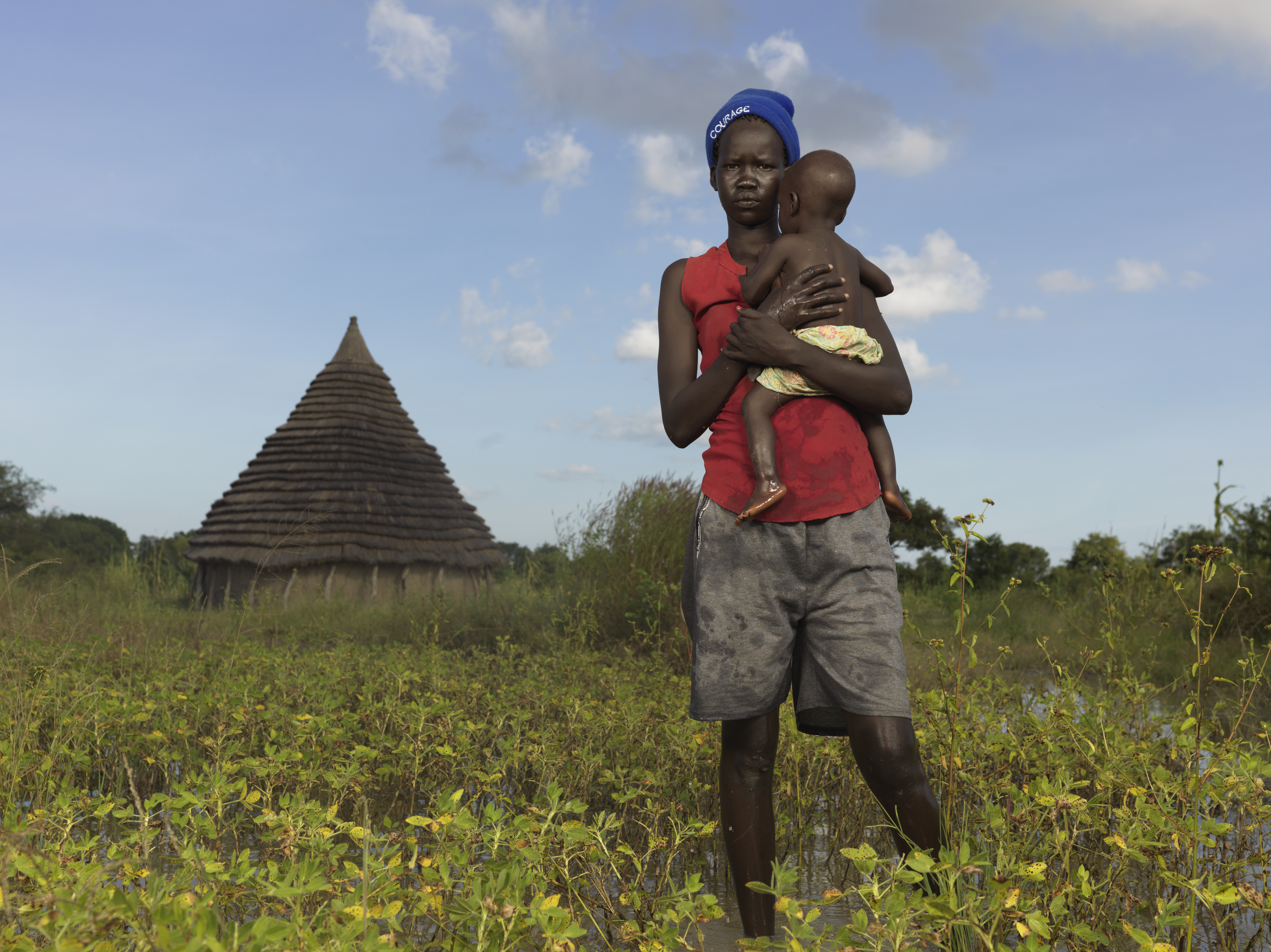 Teenage mother from South Sudan walking in a field while carrying her toddler son in her arms