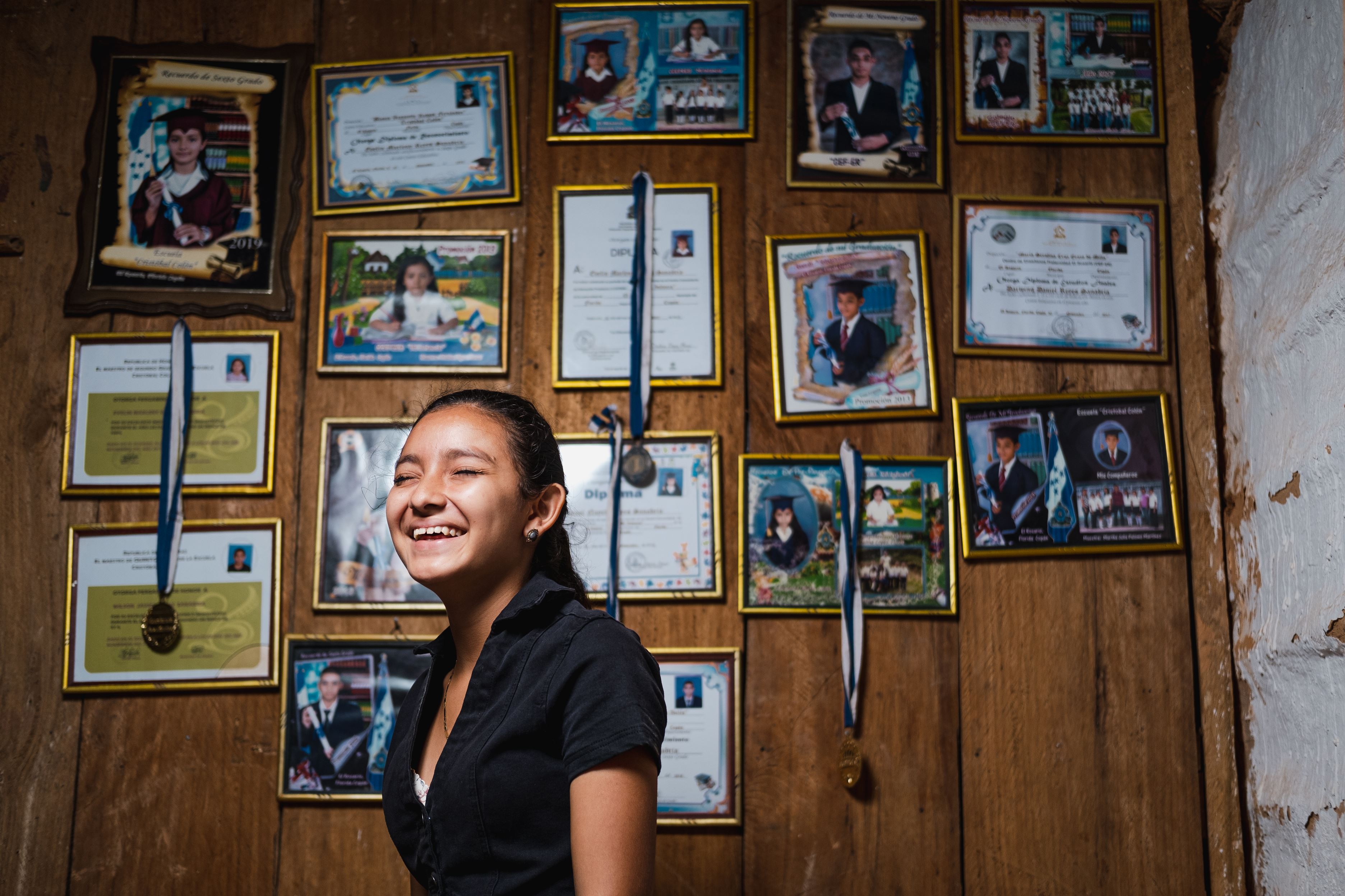 Girl from Honduras laughing while standing in front of a wall with certificates and ribbons hung on it