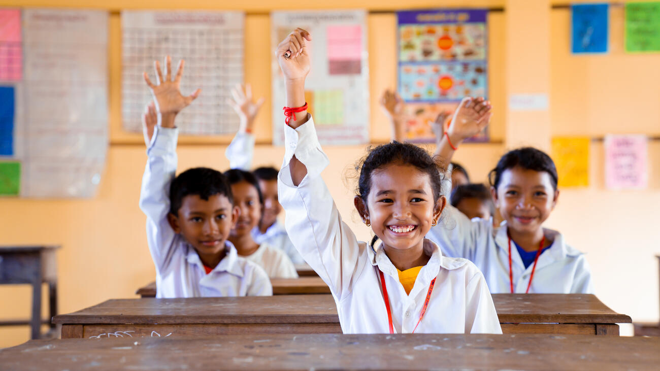 Cambodian children in a classroom, all wearing white shirts, raising their arm and smiling