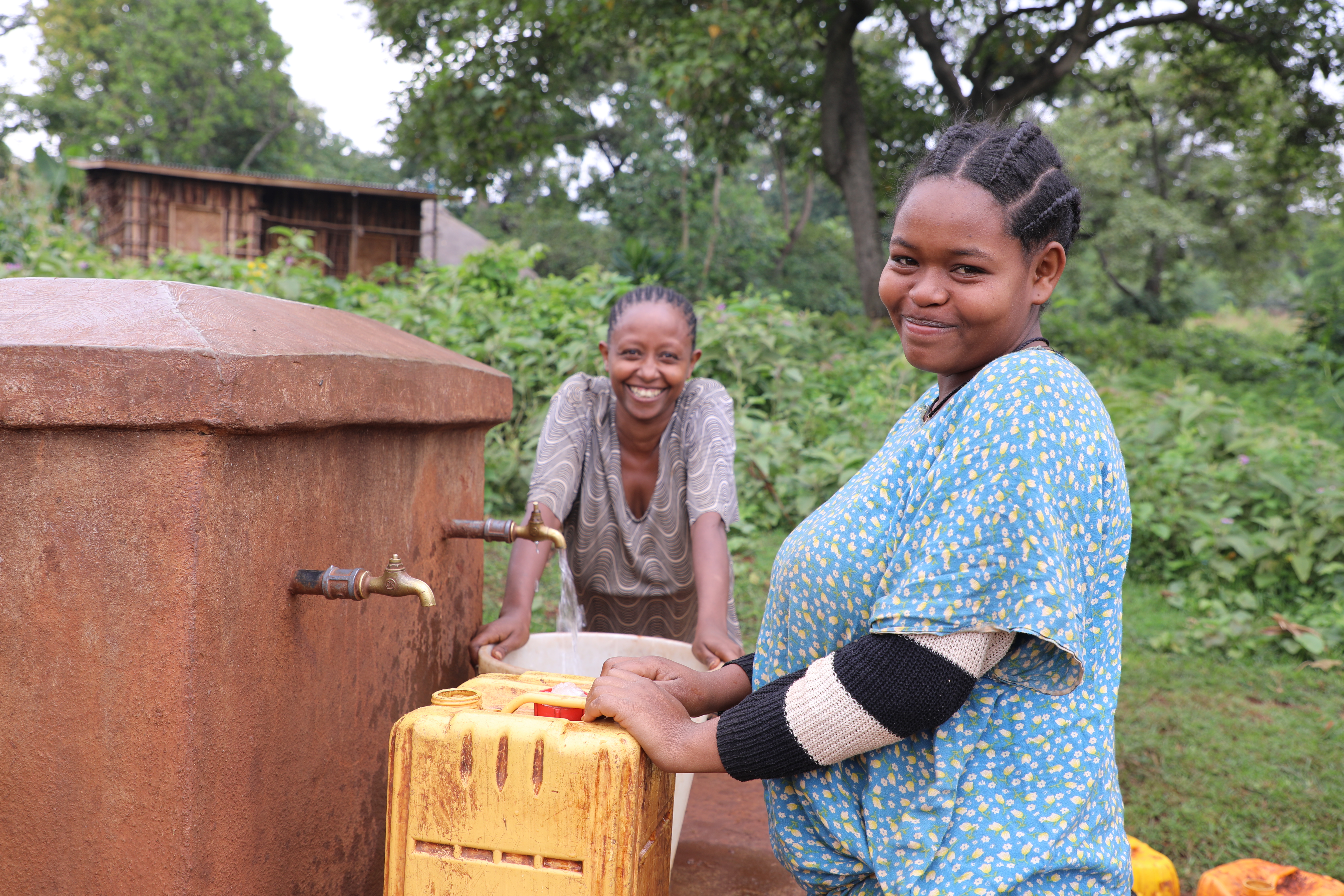 Two schoolgirls from Ethiopia filling up tubs of water for their households, while smiling and looking at the camera