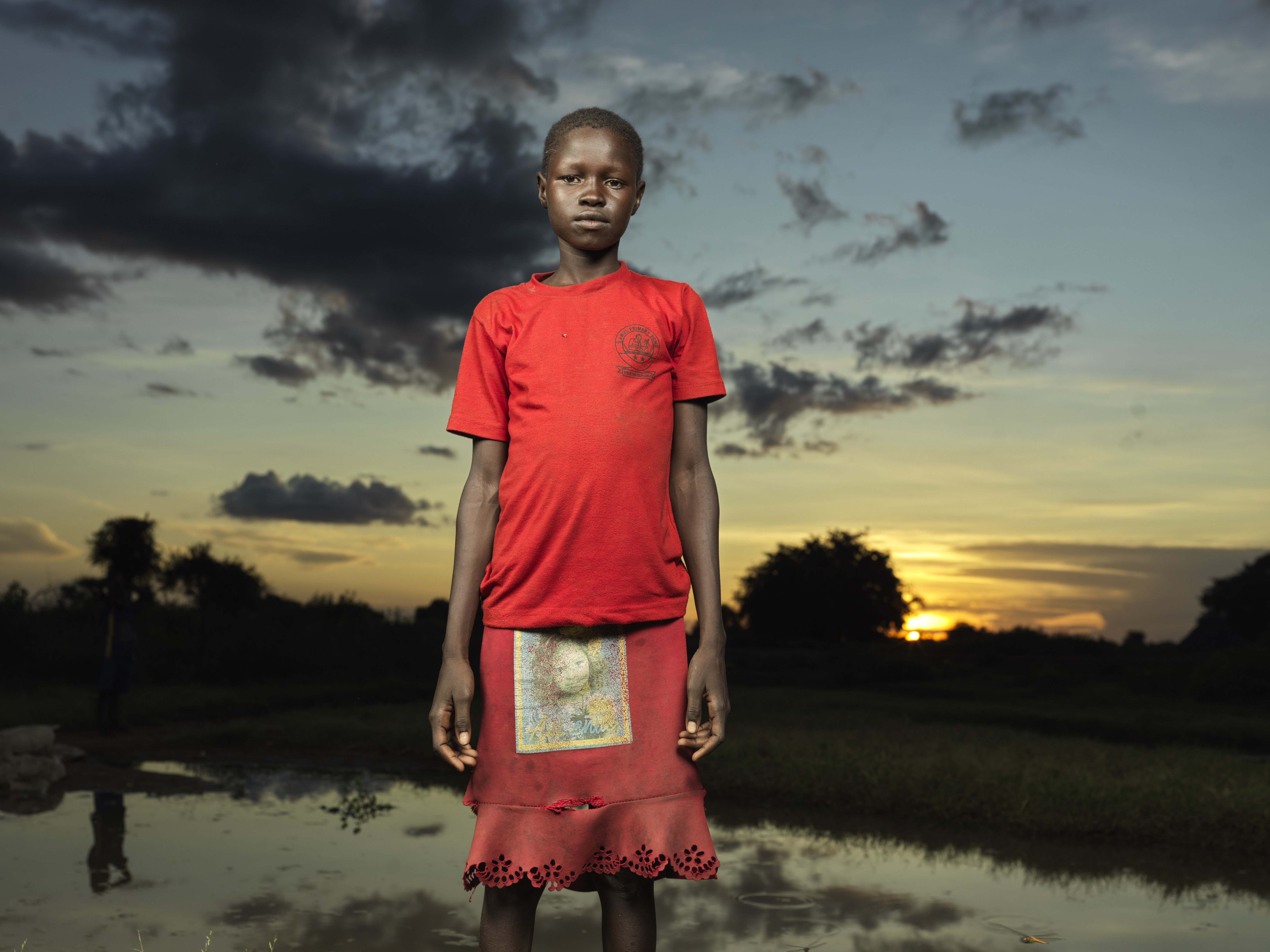 A girl form South Sudan wearing a bright red dress poses in front of a sunset
