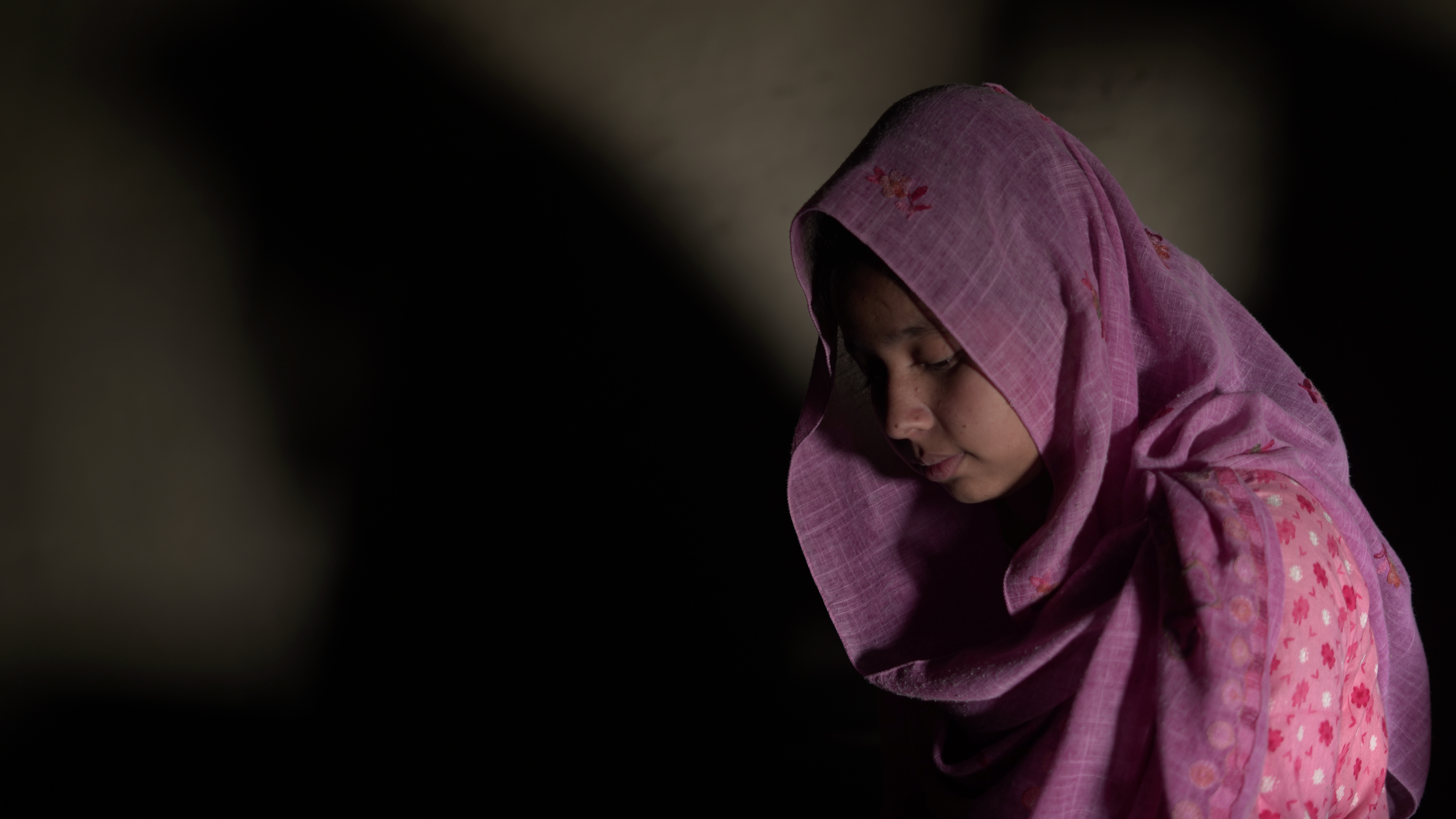 Bangladeshi girl wearing a bright pink headscarf pictured in profile, looking downwards in a dingy room