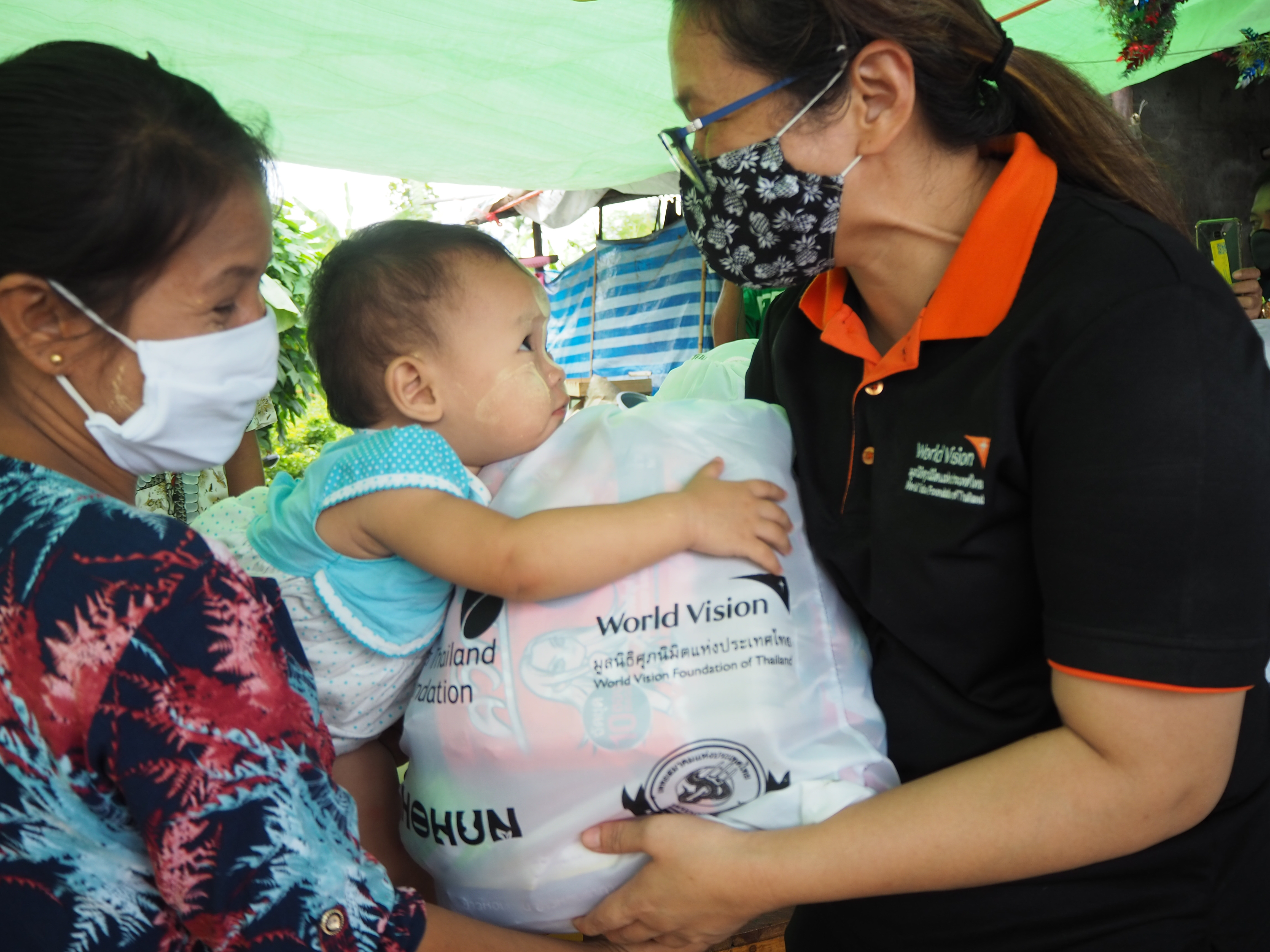 Staff member in a mask hands over a bag to support families and prevent the spread of COVID-19 in Thailand. The woman receiving the bag smiles as her child clings onto the bag