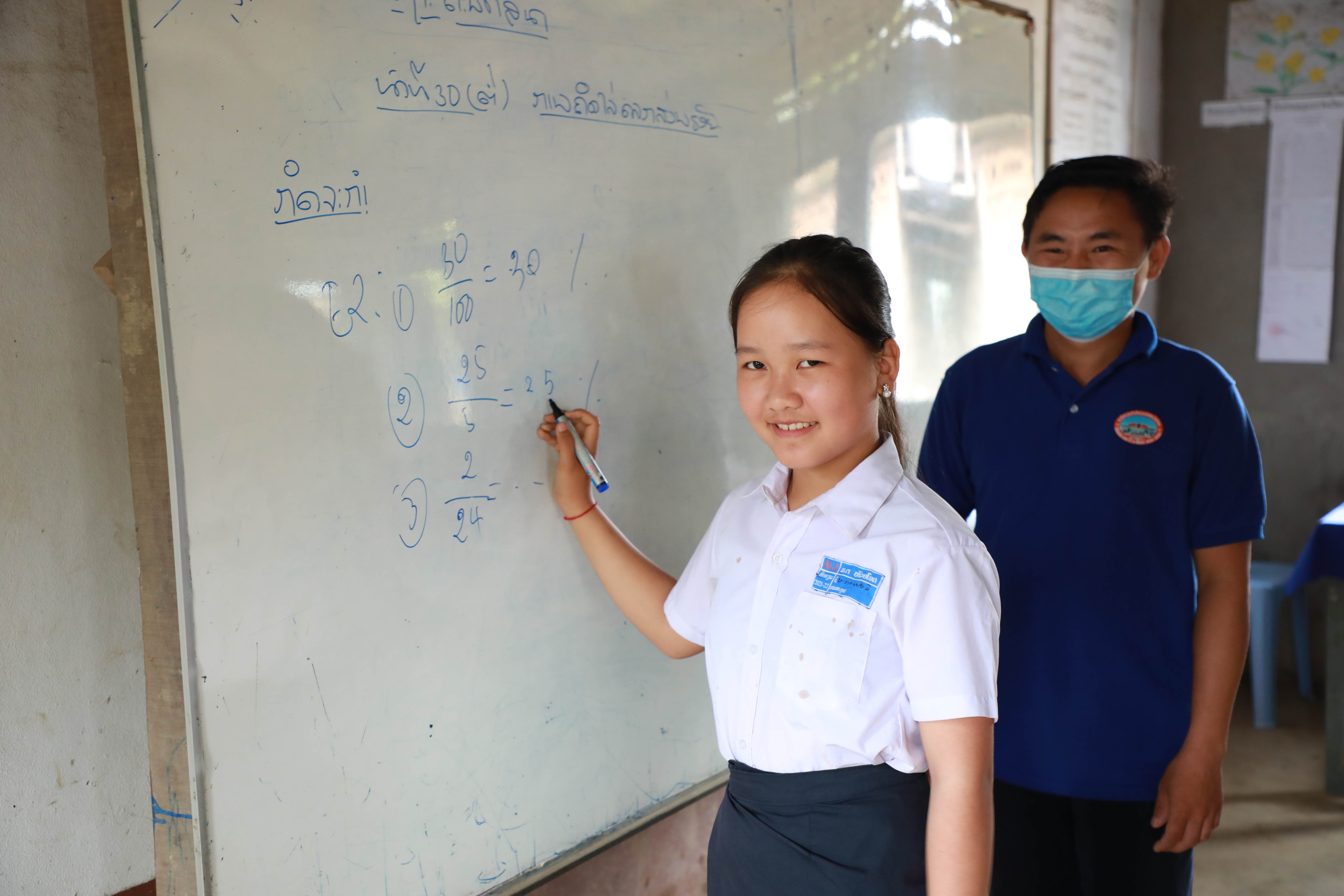 A girl student from Laos writing on a whiteboard at school while smiling at the camera. Her teacher supervises in the background while wearing a facemask.