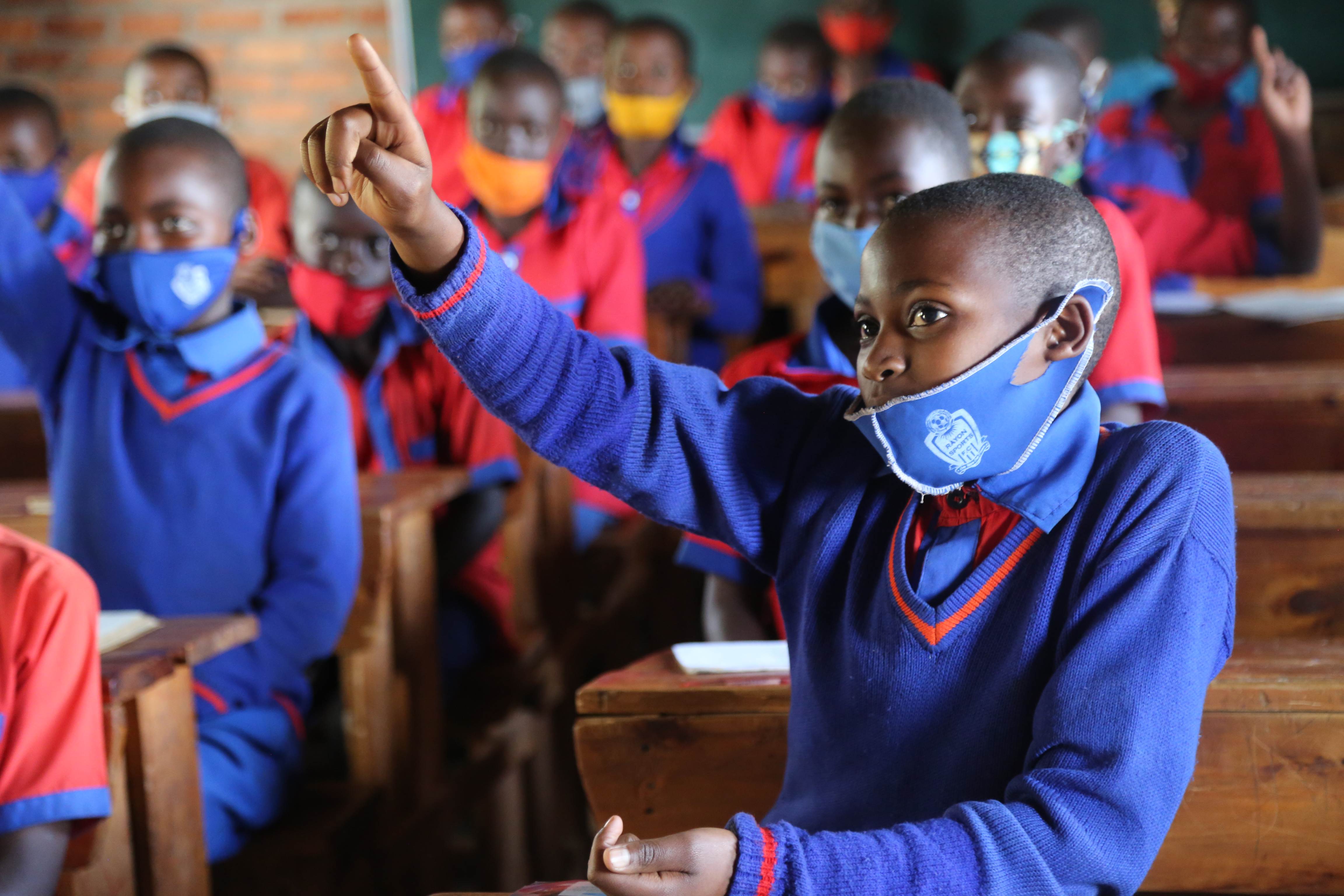 School children in blue school uniforms and facemasks in Rwanda raising their hands in class
