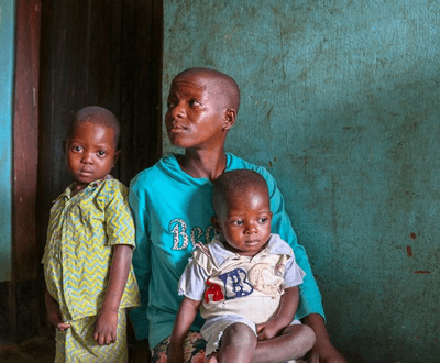 A mother in DRC looks to the side as her two children, one now recovered from malnourishment, sit on her lap