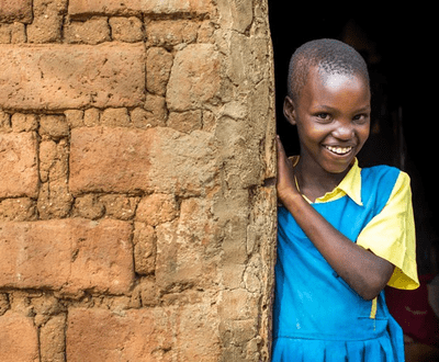 A sponsored child in Kenya wears a blue dress and poses next to a brick wall, smiling.