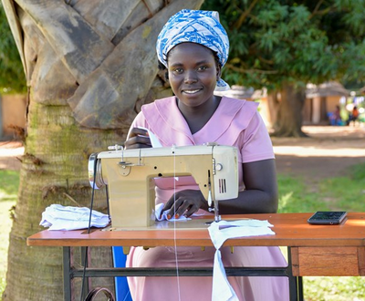Woman from South Sudan leans on a table outside in Uganda, with a sewing machine on the table