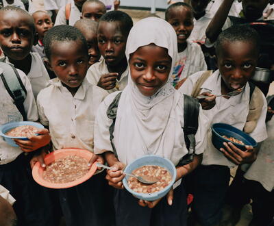 In Tanzania, 9-year-old Rukia (white hijab) holds a plate of food in front of the camera, while her classmates do the same.
