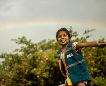Bolivian girl spreads her arms wide and smiles, with a rainbow and tree behind her
