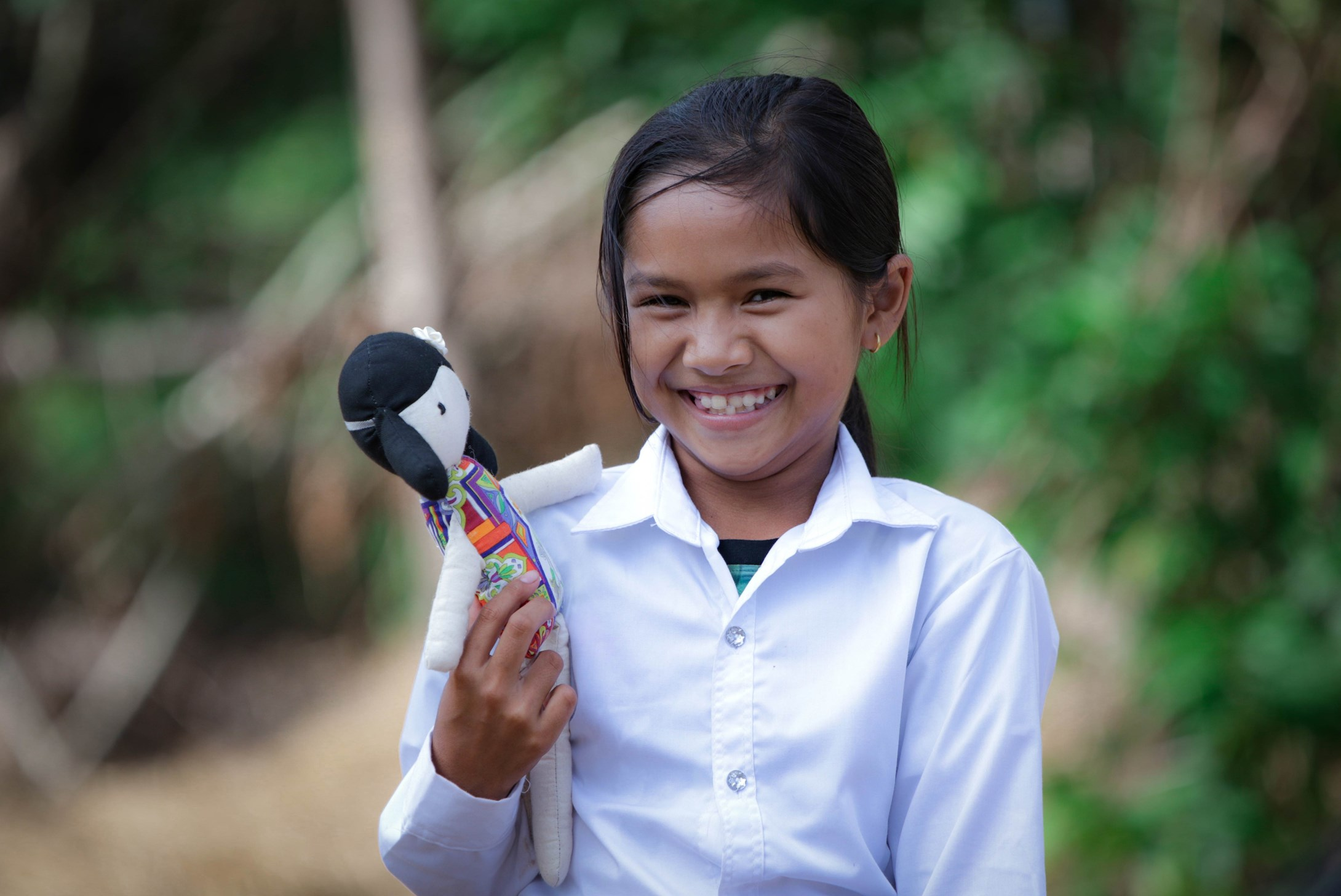 Child in Cambodia stands outside holding a doll and grinning